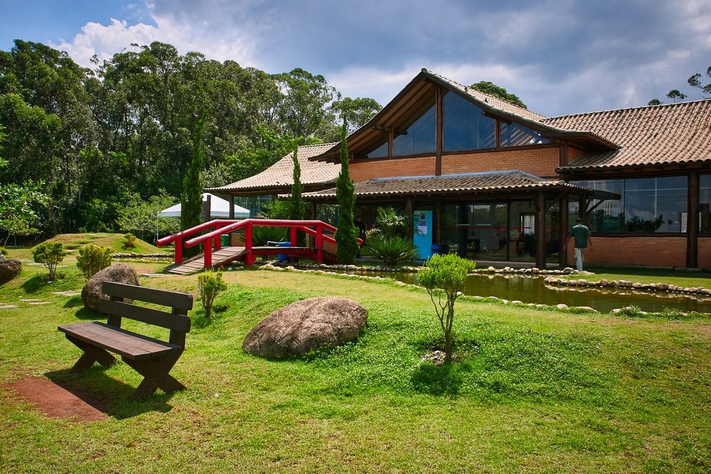 Prédio de madeira e vidro com telhado inclinado, ponte vermelha em estilo japonês e gramado com pedras e banco, no Parque Centenário da Imigração Japonesa