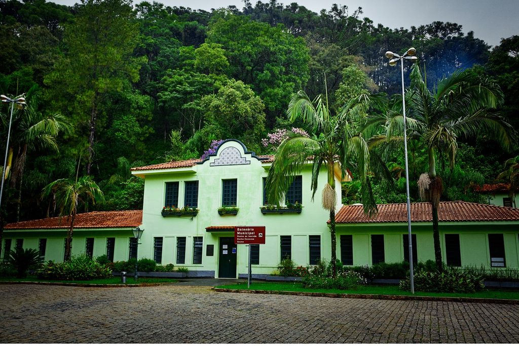 Prédio verde-claro do Balneário Municipal de Monte Alegre do Sul, em estilo neocolonial, com palmeiras e a serra coberta de mata ao fundo