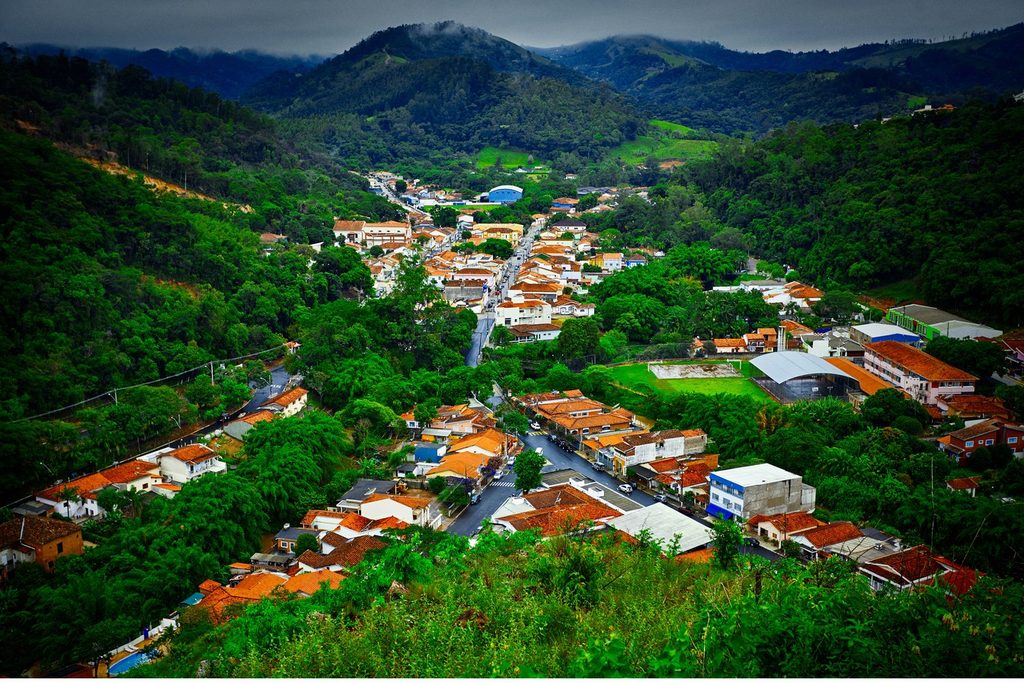 Vista do alto da cidade encaixada num vale, com casas de telhado laranja e morros cobertos de mata