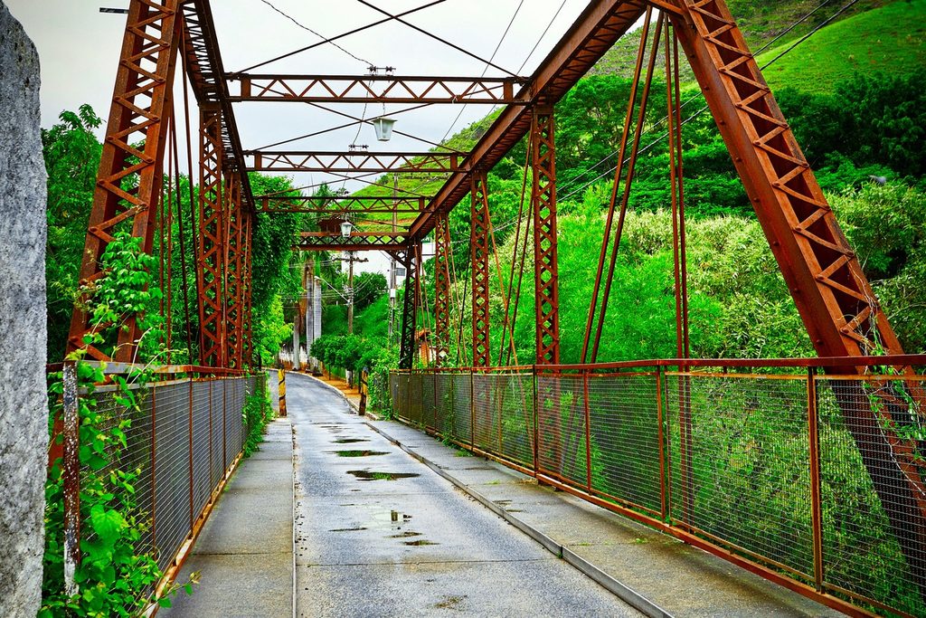 Ponte de ferro avermelhada em treliça sobre uma passagem estreita, cercada de mata