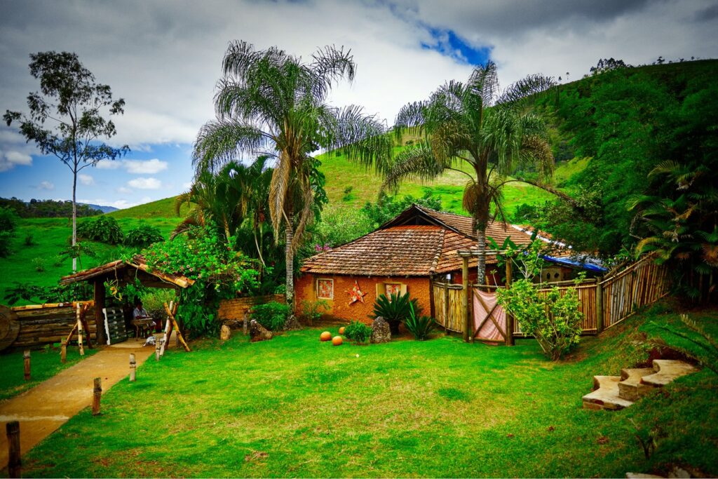 Casinha rústica de telhado de barro com varanda e cerca de madeira entre palmeiras e morros verdes, no restaurante Beira do Riacho, em Monteiro Lobato