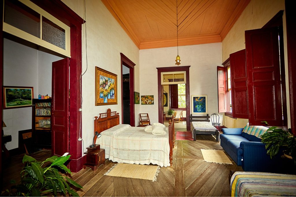 Quarto do casarão do Sítio do Picapau Amarelo, em Monteiro Lobato, com cama de madeira, portas vermelhas, forro de madeira e quadros nas paredes