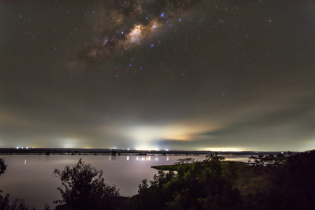 Céu noturno com a Via Láctea sobre a água e a silhueta da mata, com luzes no horizonte