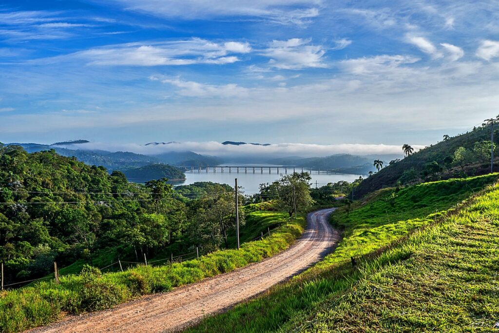 Estrada de terra entre pastos verdes descendo até a Represa Atibainha, em Nazaré Paulista, com a ponte e a serra ao fundo