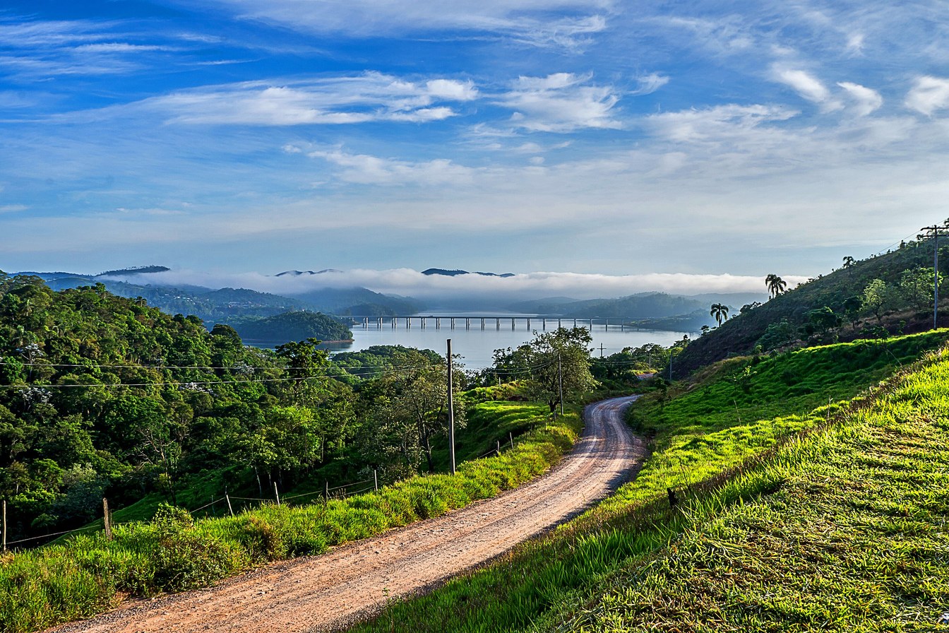 Estrada de terra entre pastos verdes descendo até a Represa Atibainha, em Nazaré Paulista, com a ponte e a serra ao fundo