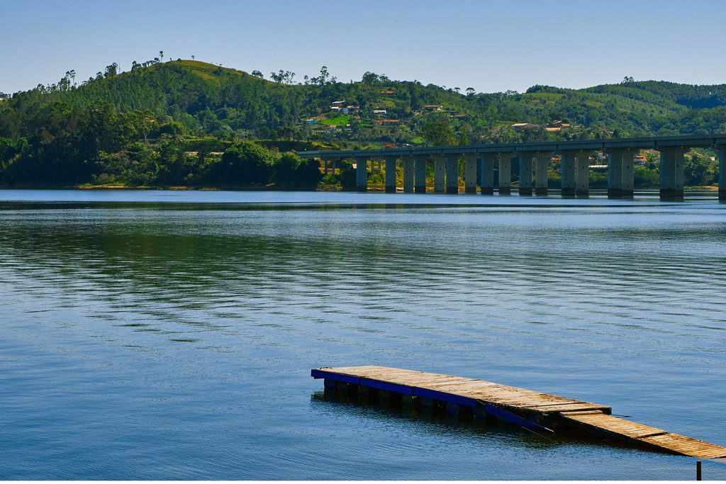 Represa Atibainha, em Nazaré Paulista, com um deque de madeira na água, a ponte e morros verdes ao fundo