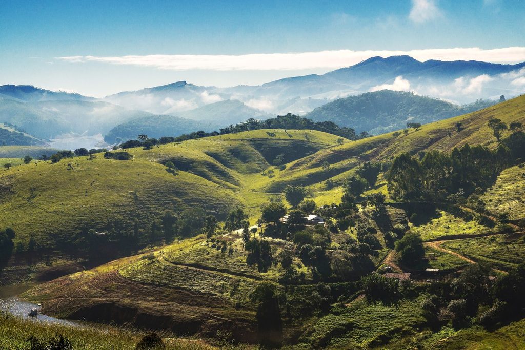 Morros verdes com chácaras e árvores, e serras com neblina ao fundo, na região da represa