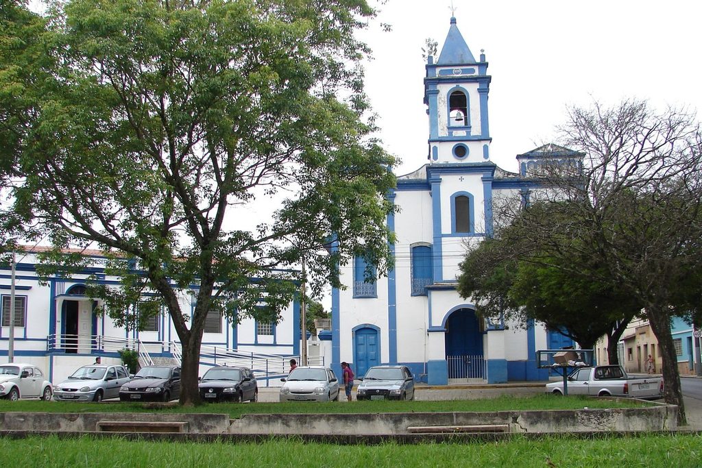 Igreja Nossa Senhora do Rosário, branca com detalhes azuis, ao lado do prédio da Santa Casa, atrás de árvores