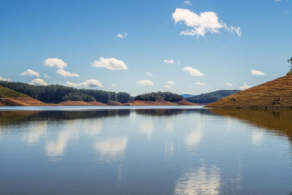Água calma da represa de Paraibuna refletindo nuvens brancas e o céu azul, com margens de terra e mata
