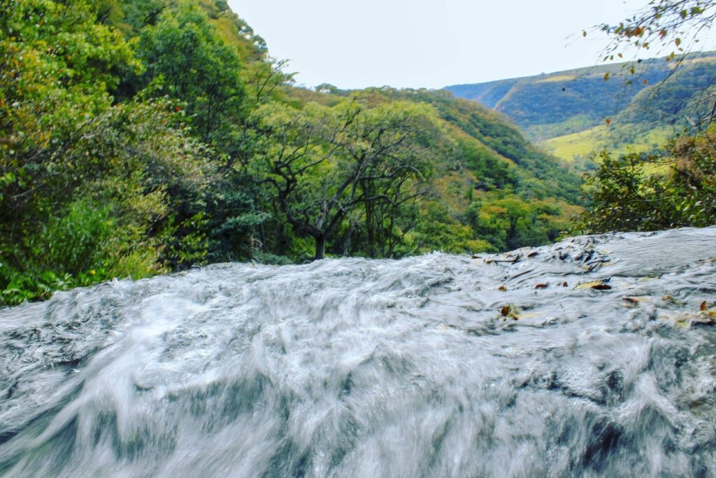 Água correndo forte sobre a pedra no alto de uma cachoeira, com o vale verde e os morros de Pedregulho ao fundo