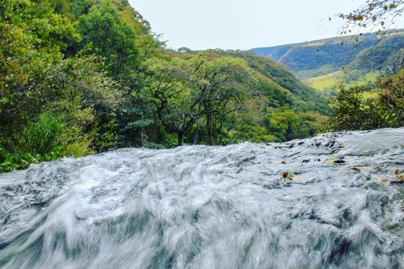 Água correndo forte sobre a pedra no alto de uma cachoeira, com o vale verde e os morros de Pedregulho ao fundo