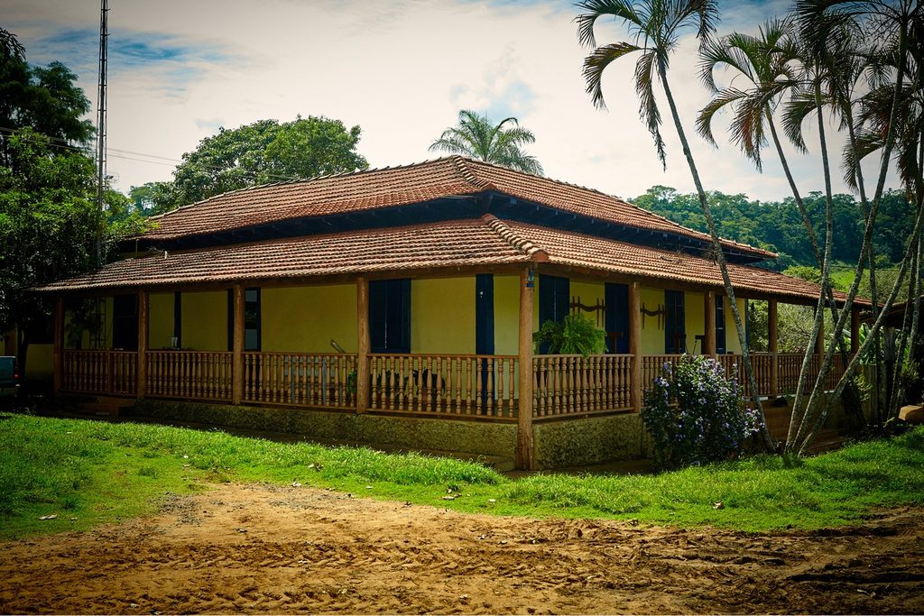 Casarão amarelo de fazenda com varanda de madeira e telhado de barro entre palmeiras