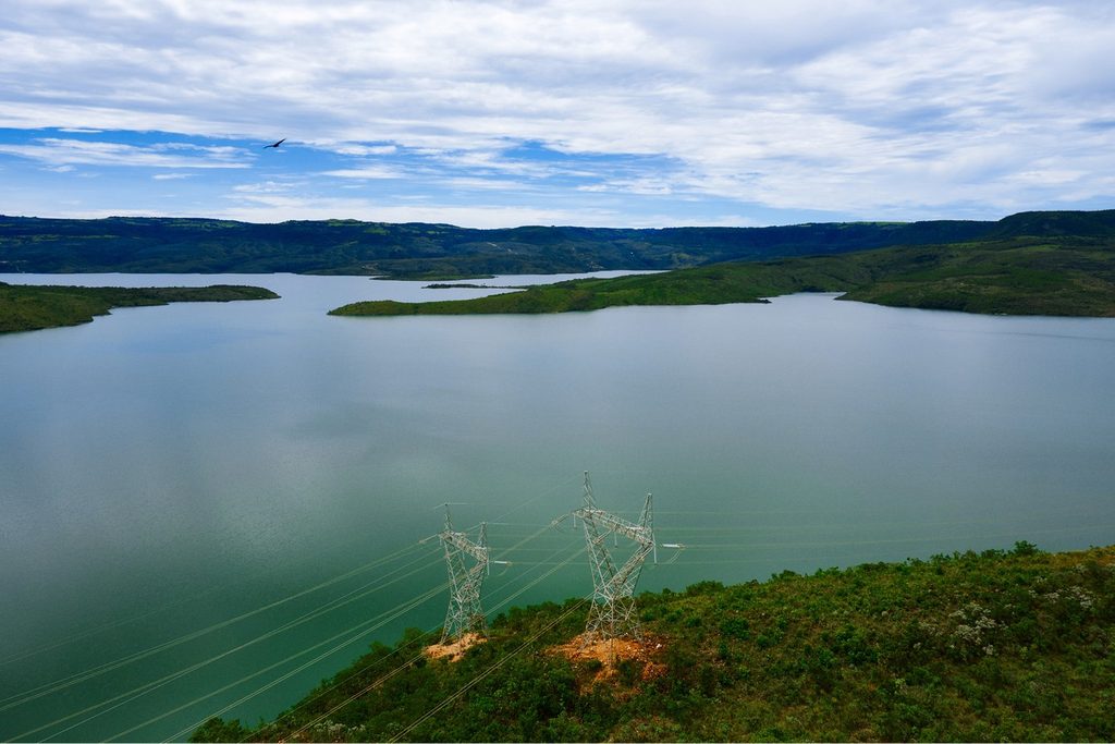 Lago extenso de água verde entre morros e chapadas, com torres de transmissão na margem