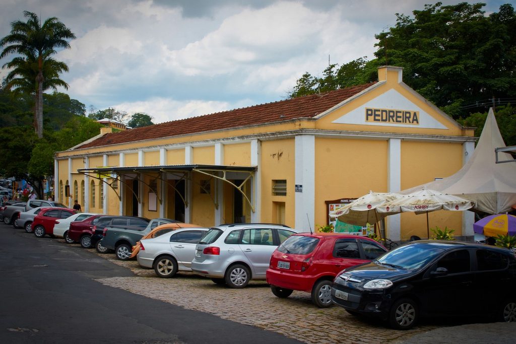 Antiga estação de trem amarela com a placa Pedreira no alto, toldos de feira e carros estacionados na frente