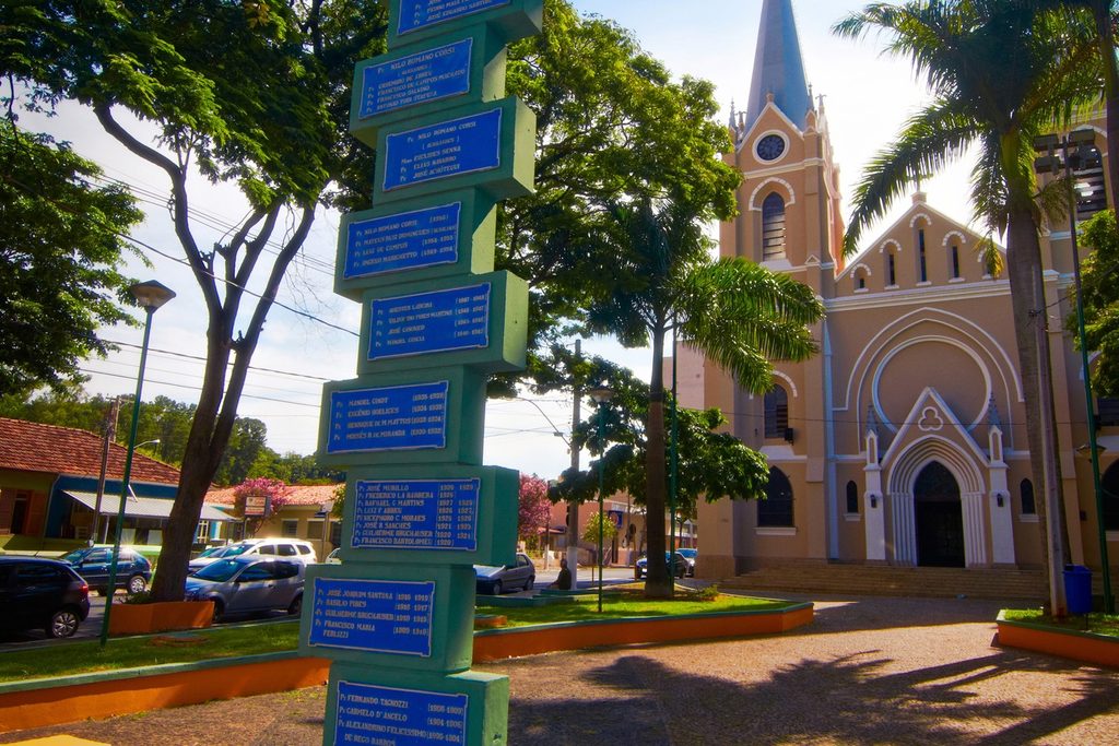 Fachada neogótica rosa-clara da Igreja Matriz de Sant'Ana, com torre pontiaguda e janelas em arco, atrás de uma praça com palmeiras e um totem de placas azuis