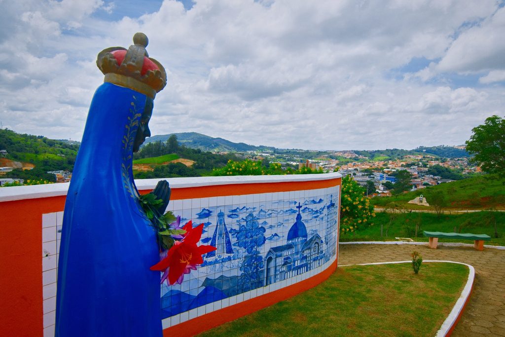 Imagem de Nossa Senhora Aparecida em azul diante de painel de azulejos pintados, com a cidade de Pedreira e os morros ao fundo
