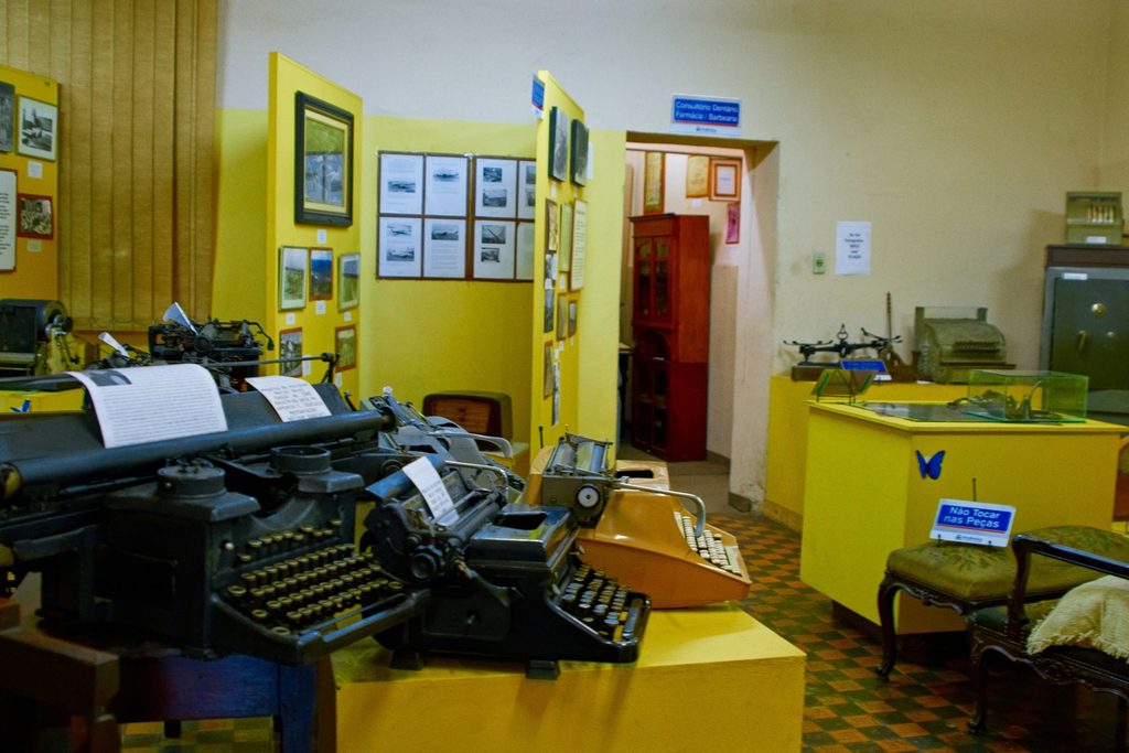 Sala de museu com paredes amarelas, máquinas de escrever antigas e vitrines com objetos e fotografias antigas