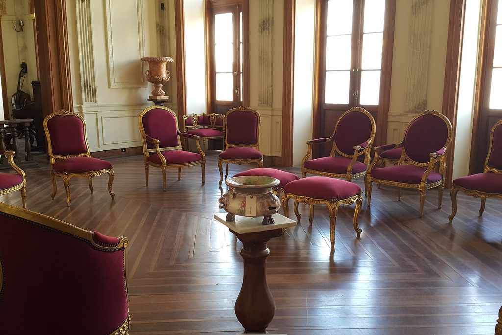 Sala do Palacete Palmeira, em Pindamonhangaba, com cadeiras douradas estofadas em vermelho, vasos e piso de madeira