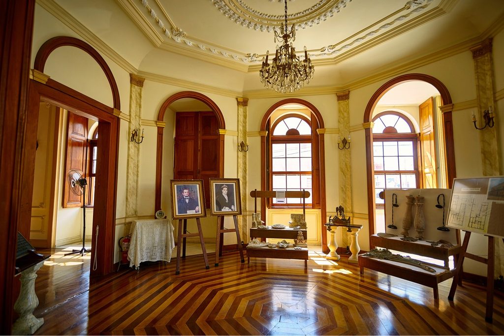 Salão do museu com piso de tacos desenhados, lustre de cristal, janelas em arco e retratos antigos em cavaletes