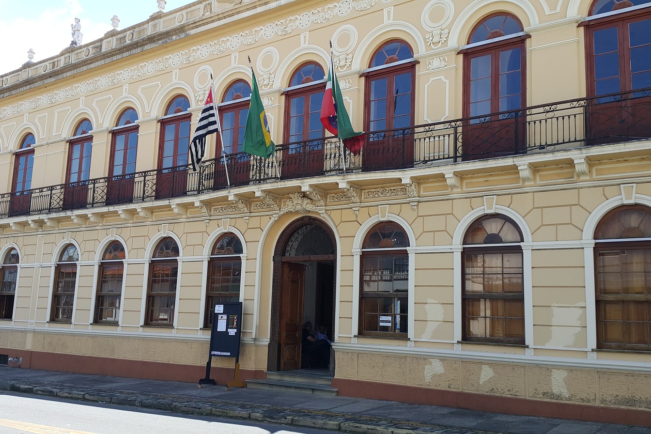 Fachada amarela do Palacete Palmeira, em Pindamonhangaba, com janelas em arco, sacada de ferro e bandeiras na entrada