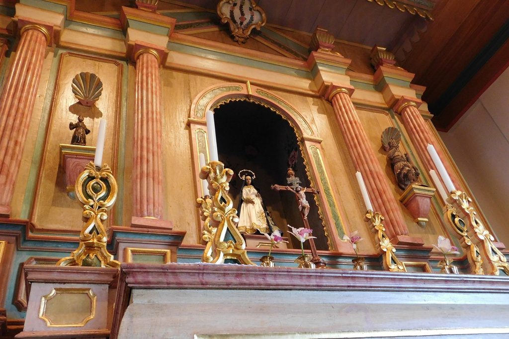Altar de madeira entalhada e pintada em tons de rosa e dourado, com imagens de santos e um crucifixo