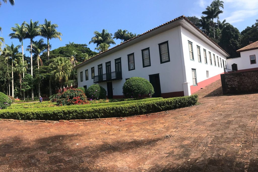 Casa-grande da Fazenda do Pinhal, branca, de dois pavimentos, com janelas escuras, jardim e palmeiras ao fundo
