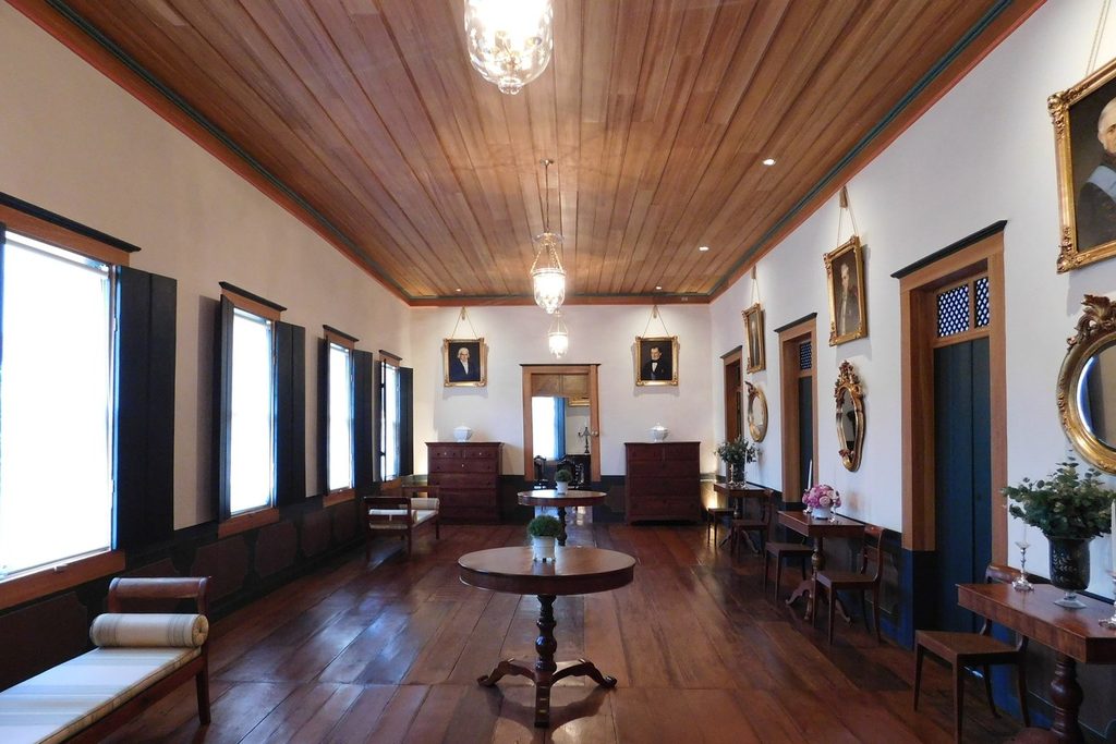 Salão comprido da casa-sede com assoalho e forro de madeira, retratos antigos nas paredes e móveis de época