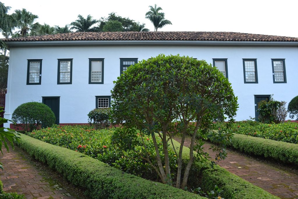 Fachada branca da casa-sede da Fazenda do Pinhal com janelas verdes e jardim de buxinhos em canteiros