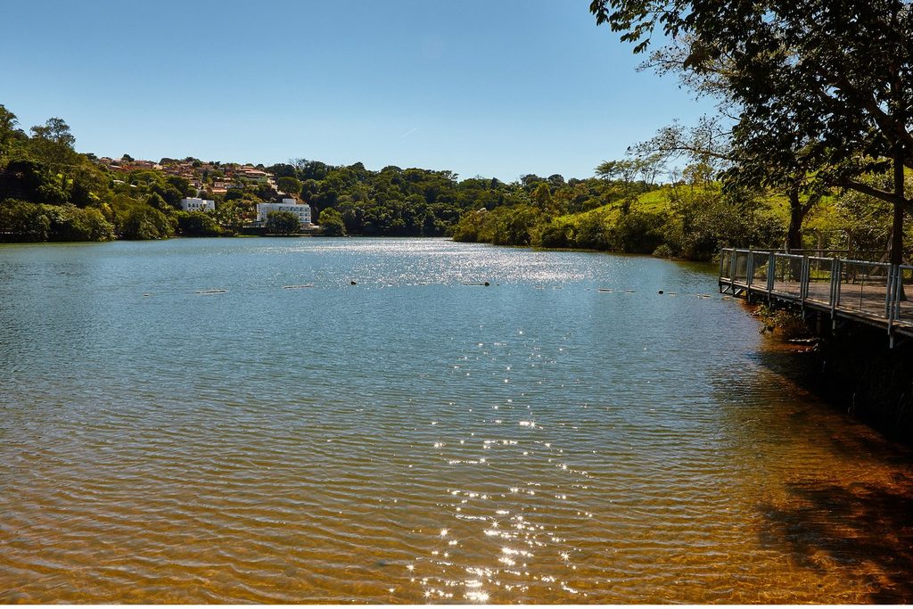 Represa de água calma com reflexos do sol, píer de madeira na margem e casas entre as árvores