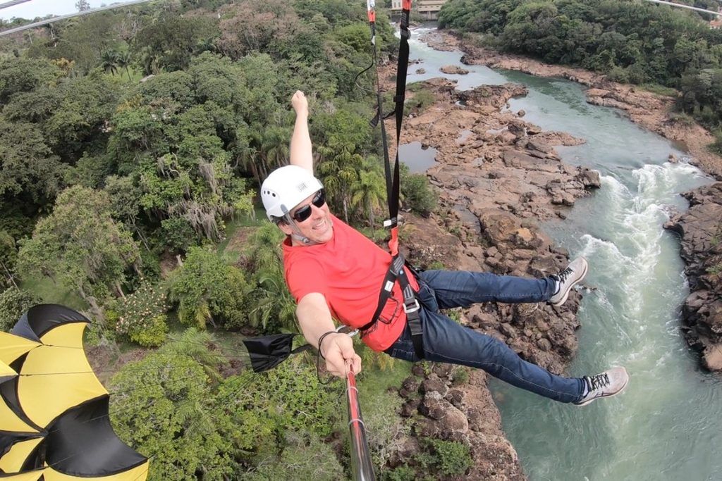 Pessoa de capacete descendo pela tirolesa no alto, com o rio correndo entre pedras e mata lá embaixo