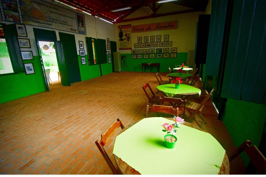 Salão da Casa do Samba com mesas de toalha verde, chão de tijolos e fotos antigas de sambistas nas paredes