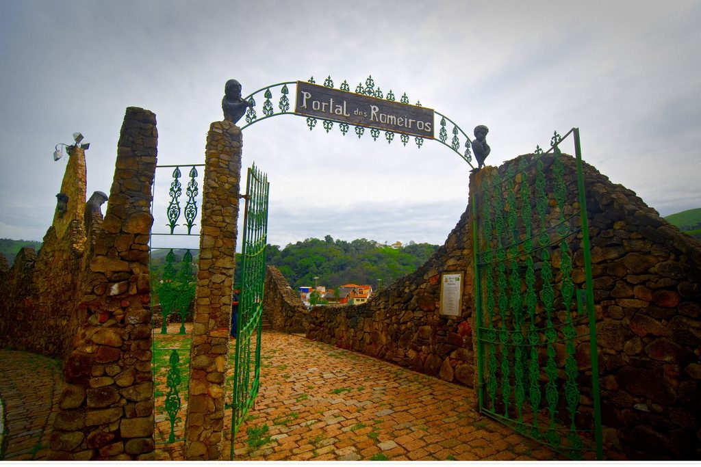 Portal de pedra com arco de ferro e a inscrição Portal dos Romeiros, com um caminho de paralelepípedos