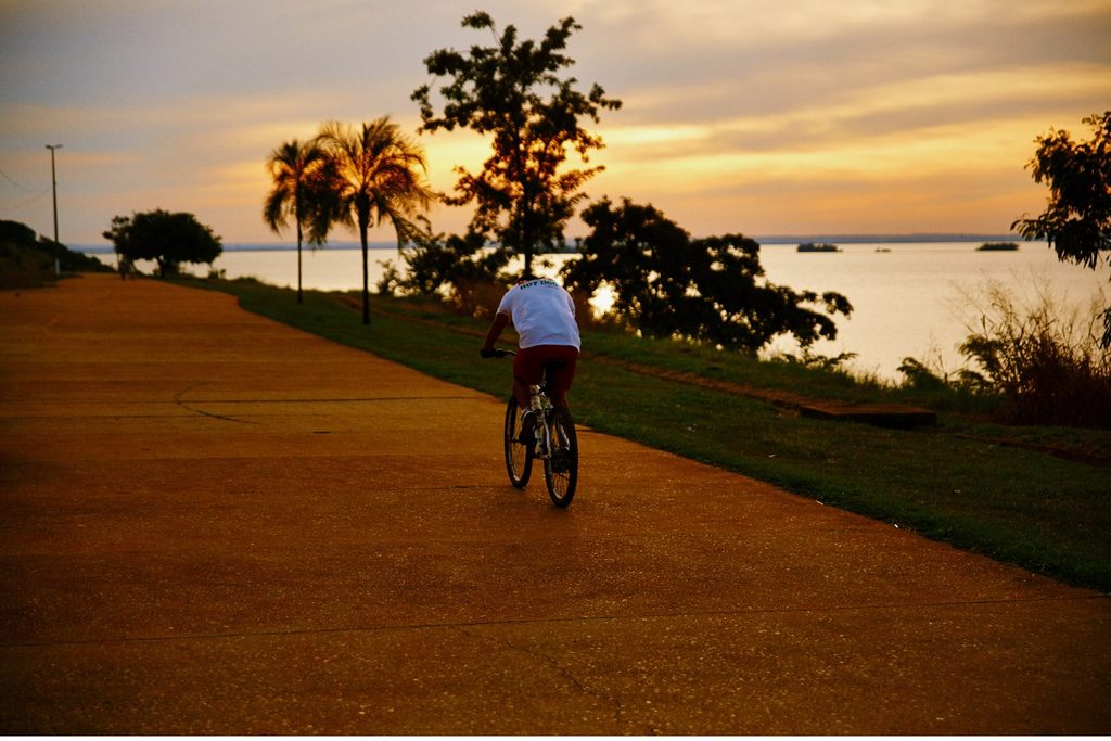 Ciclista pedalando na pista da orla fluvial de Presidente Epitácio ao entardecer, com palmeiras e o rio ao fundo