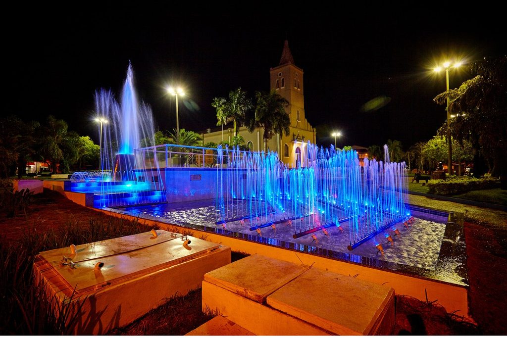 Fonte luminosa com jatos azuis iluminados à noite na Praça da Matriz de Presidente Epitácio, com a igreja ao fundo