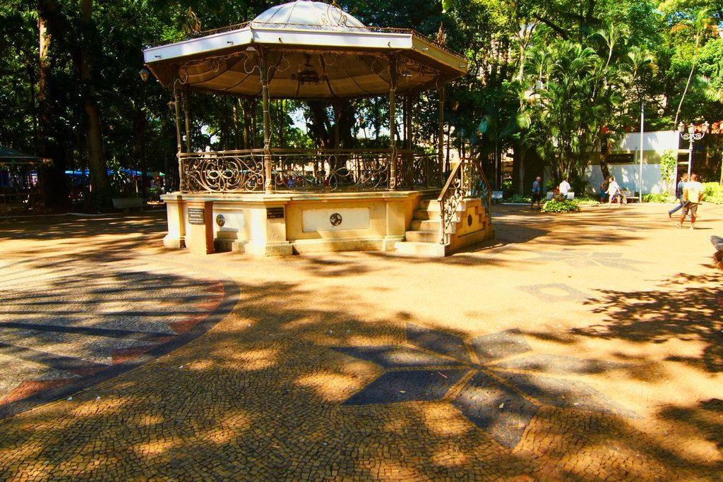 Coreto de ferro com cúpula, sobre base de pedra, numa praça arborizada com piso de pedras portuguesas