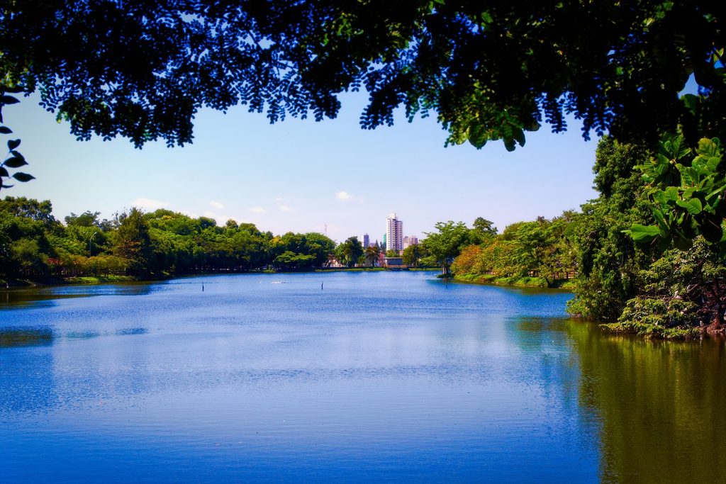 Lago de água azul cercado de árvores, com prédios da cidade ao fundo, no Parque Lago Azul