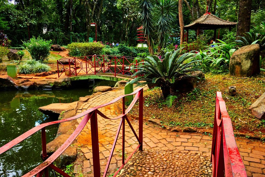 Jardim japonês com ponte de corrimão vermelho sobre um lago, caminho de pedras, plantas tropicais e quiosque com telhado oriental