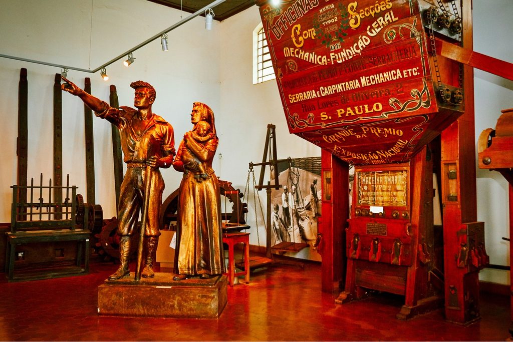 Sala do Museu do Café com estátua de casal de imigrantes, máquinas antigas de beneficiar café e uma grande caixa vermelha com letreiros