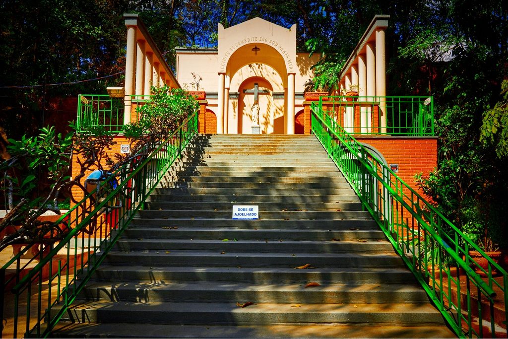 Escadaria com corrimões verdes subindo até uma capela clara com colunas no Santuário das Sete Capelas