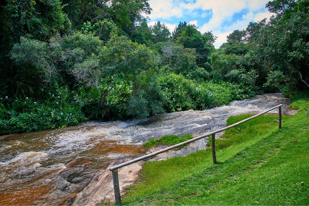 Riacho com corredeira sobre pedras, entre mata fechada e gramado, na área do Casarão do Café, em Salesópolis