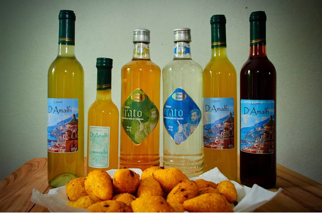 Garrafas de cachaça artesanal de várias cores ao lado de uma cesta de bolinhos fritos