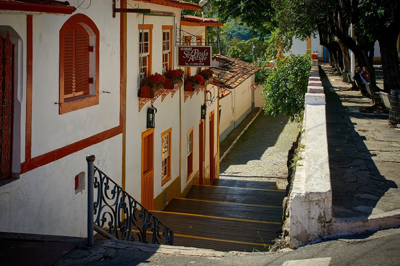 Rua em ladeira com escadaria de pedra, casarões coloniais brancos de janelas vermelhas e floreiras, no centro histórico de Santana de Parnaíba