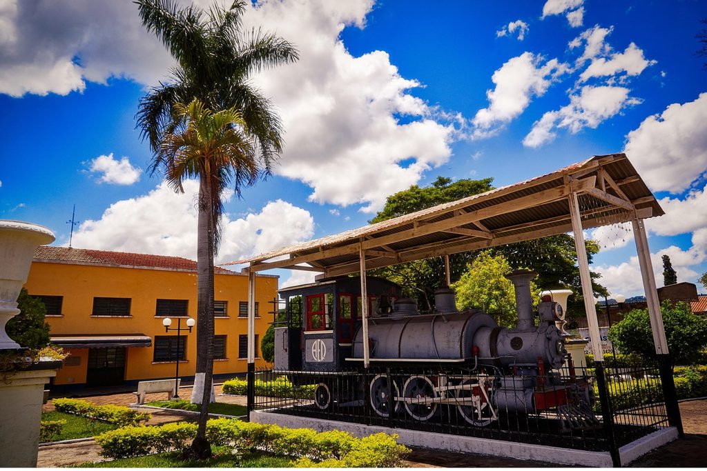 Locomotiva a vapor preservada sob cobertura, com palmeira e casa amarela ao fundo