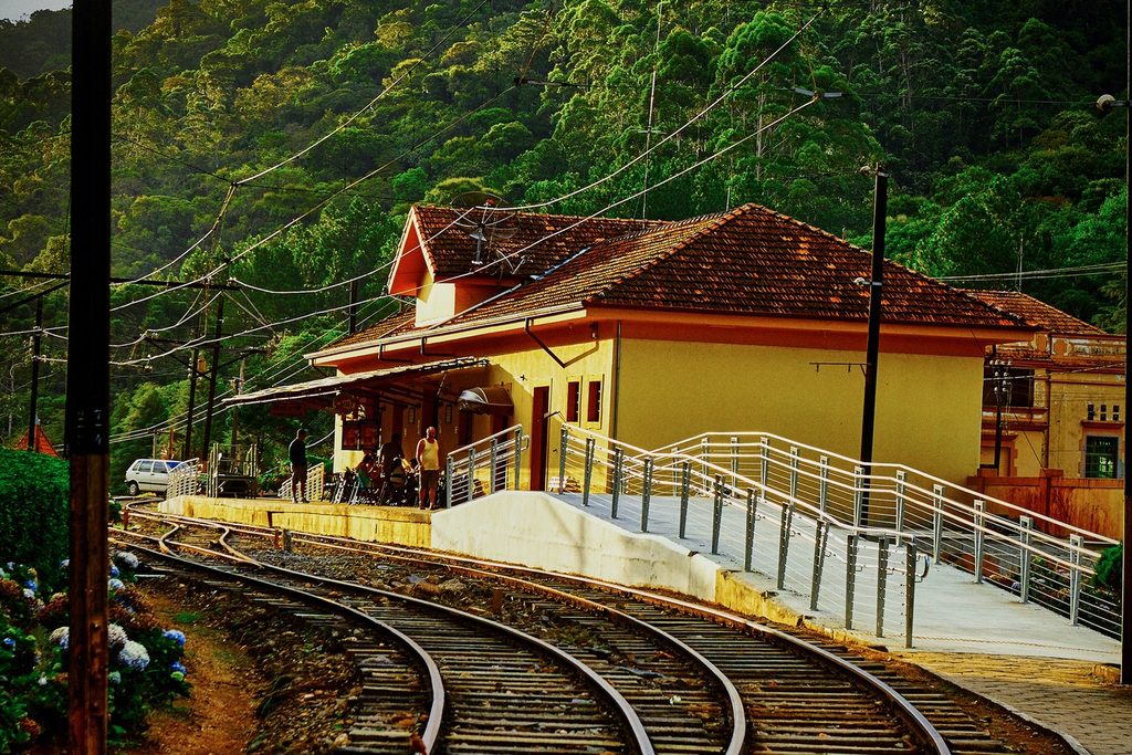 Estação ferroviária amarela de telhado vermelho com plataforma, trilhos em curva e mata ao fundo, a Estação Eugênio Lefèvre