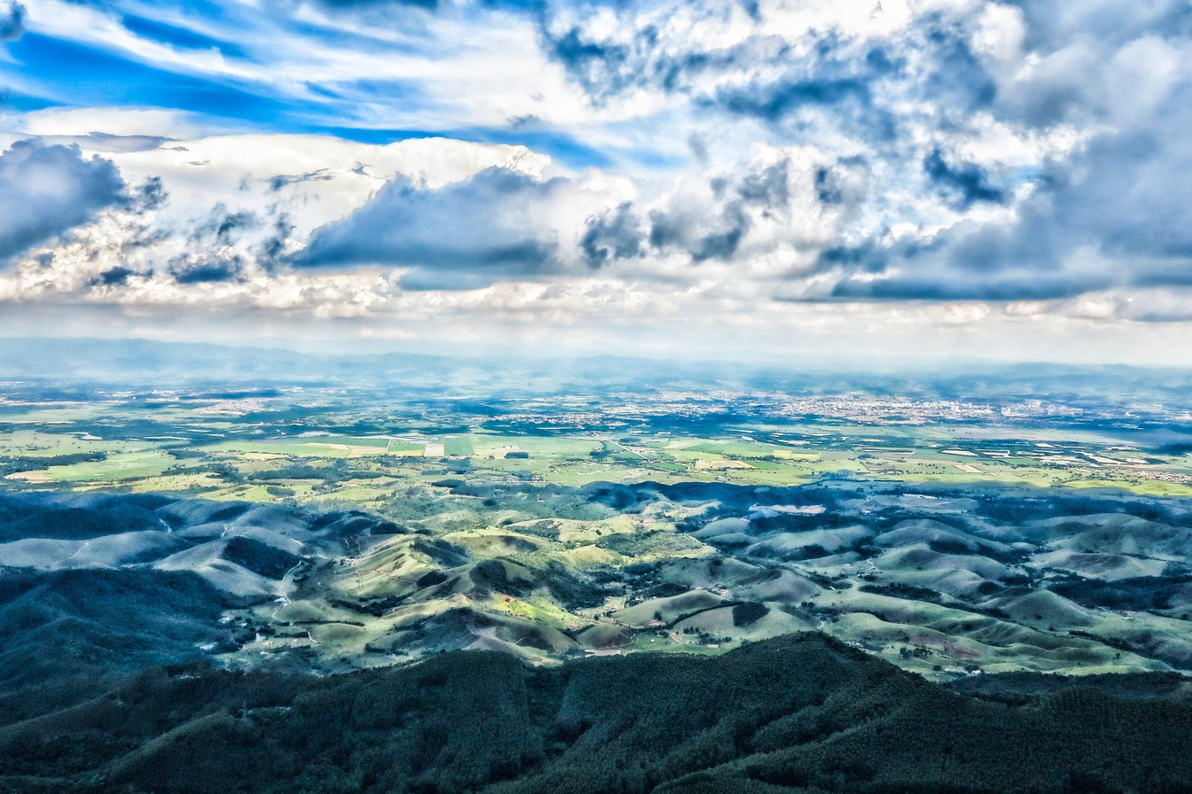 Vista do alto do Pico Agudo sobre o vale, com morros verdes, nuvens baixas e céu azul com nuvens até o horizonte