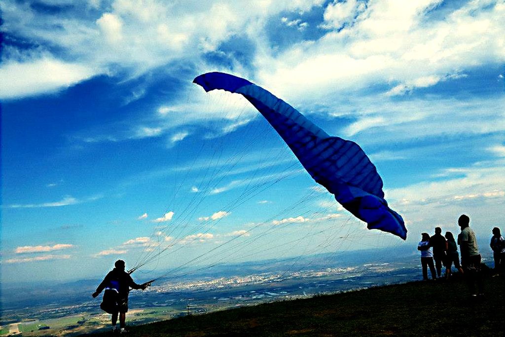 Piloto de parapente se preparando para decolar com a vela azul inflada no alto do Pico Agudo, observado por outras pessoas