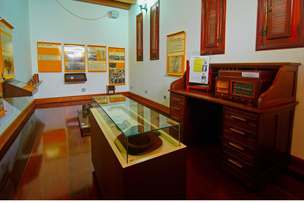 Sala do Museu da Imigração de Santa Bárbara d'Oeste com vitrine, escrivaninha antiga de madeira e painéis com fotos históricas