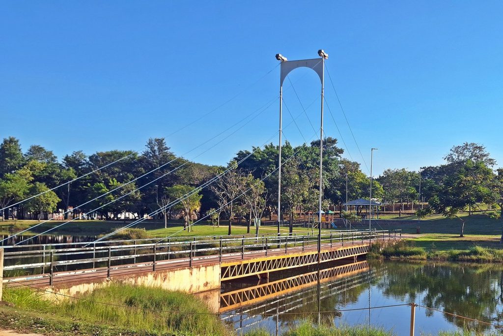 Passarela suspensa sobre o lago do Parque Araçariguama, com gramado e árvores na outra margem