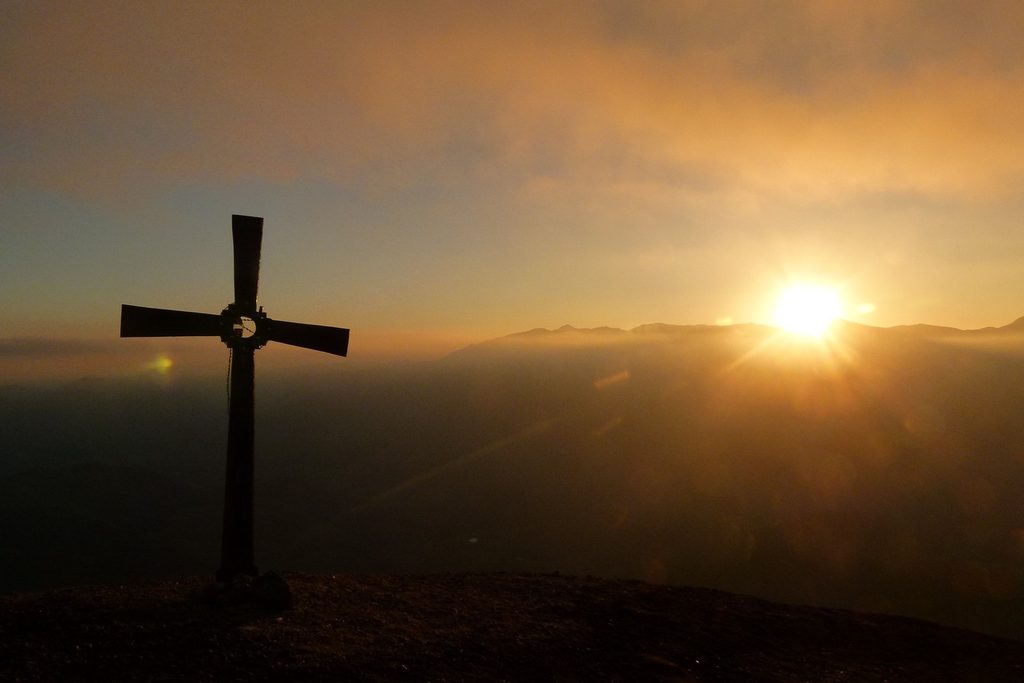 Silhueta de uma cruz no alto de um morro diante do sol se pondo sobre as montanhas, com céu alaranjado