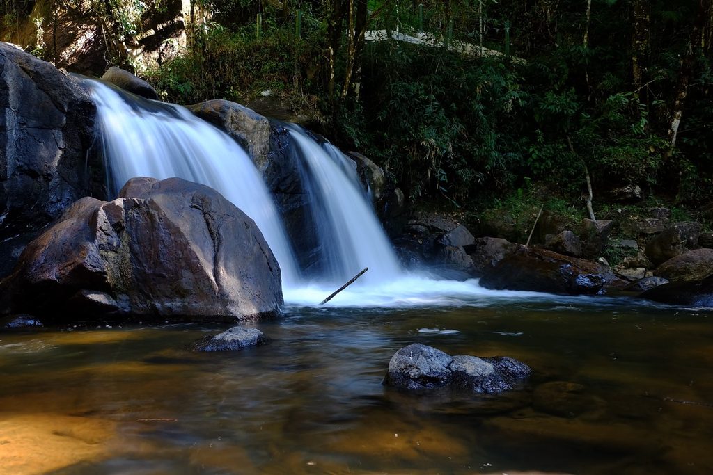 Cachoeira Pedro David descendo em duas quedas brancas entre grandes pedras escuras, sobre um poço de água cor de chá, em São Francisco Xavier